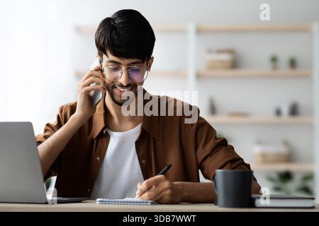 Jeune homme souriant tout en parlant au téléphone et en prenant des notes dans un espace de travail moderne Banque D'Images