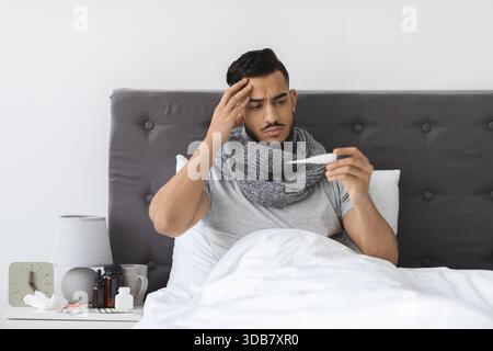 Homme malade du moyen-Orient assis dans le lit tenant le thermomètre et se sentant malheureux avec des symptômes de grippe Banque D'Images