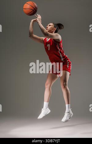 Femme dans l'équipement de basket rouge tirant la balle à mi-saut dans la lumière de studio. Athlétisme, confiance et action figés dans le mouvement. Banque D'Images