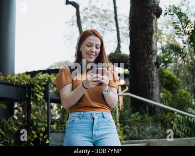 Jeune femme heureuse utilisant son smartphone à l'extérieur dans un environnement vert urbain. Concept moderne de style de vie, de mobilité et de technologie. Banque D'Images