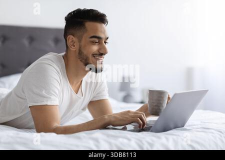 Bel homme arabe se détend dans le lit, apprécie le café tout en naviguant sur Internet Banque D'Images