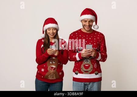 Couple portant des pulls et des chapeaux de Noël profitant de leurs smartphones pendant la saison des fêtes Banque D'Images