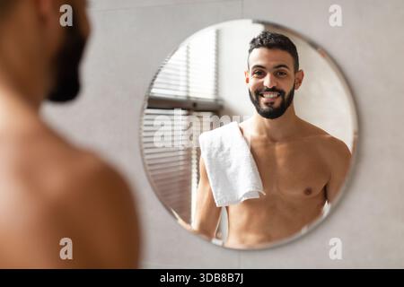 Confiant jeune homme arabe barbu se regardant dans un miroir dans une salle de bain moderne Banque D'Images