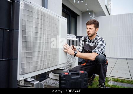 Technicien effectuant l'entretien de la pompe à chaleur extérieure près de l'extérieur du bâtiment résidentiel. Homme installant la pompe à chaleur de source d'air, à l'aide d'outils. Conc Banque D'Images