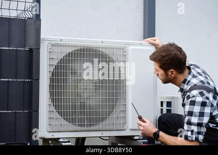 Technicien effectuant l'entretien de la pompe à chaleur extérieure près de l'extérieur du bâtiment résidentiel. Homme installant la pompe à chaleur de source d'air, à l'aide d'outils. Conc Banque D'Images