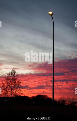 le lampadaire silhouetted se dresse haut contre un ciel de coucher de soleil spectaculaire avec des teintes rouges et oranges vibrantes, contrastant avec les arbres silhouettés et un calme, Banque D'Images