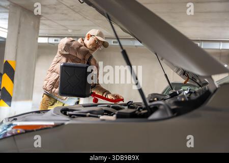 Homme versant du liquide lave-glace sous le capot d'une voiture. Entretien régulier de la voiture dans un parking ouvert. Banque D'Images
