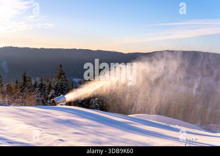 Le canon à neige fonctionne sur une piste de ski pour produire de la neige artificielle. Le soleil brille sur le paysage montagneux tandis que la neige s’accumule pour les activités de sports d’hiver. Banque D'Images
