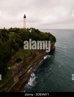 Vue aérienne du phare blanc et dur debout contre le ciel orageux, contrastant avec la côte accidentée et la mer bleu profond, Biarritz, Banque D'Images