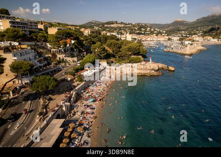 Vue aérienne de la plage ensoleillée vivante avec des parasols vibrants et des baigneurs, contrastée par les eaux turquoises calmes et le port lointain, Cassis, Prov Banque D'Images