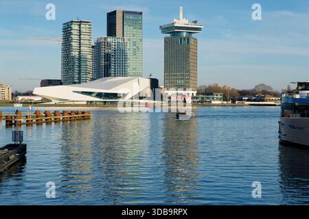 De grands bâtiments s'élèvent contre un ciel bleu, leurs reflets scintillant sur l'eau calme en contrebas. Un petit bateau se déplace à travers la scène, ajoutant de la vie au Banque D'Images