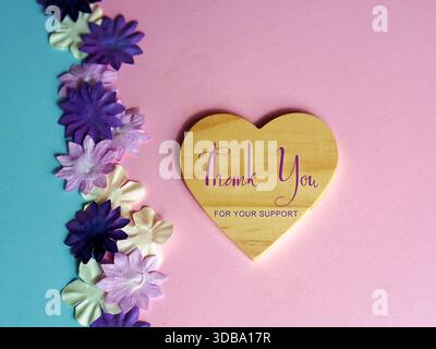 Merci de votre soutien. Mots écrits sur un coeur en bois sur un fond rose avec des fleurs violettes. Banque D'Images