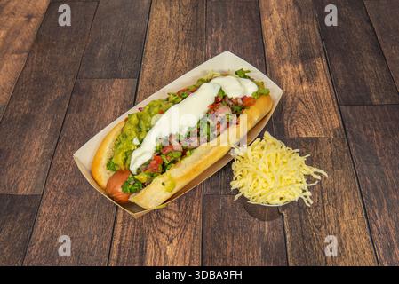 Un hot-dog mexicain avec une saucisse enveloppée dans du bacon, des haricots frits, du guacamole, du ​​jalapeños tranché, de la crème sure, et du cheddar râpé Banque D'Images