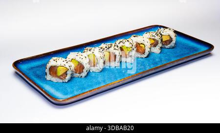 Rouleaux de sushi classiques remplis de saumon et d'avocat, enrobés de riz et de graines de sésame, servis sur une plaque de céramique bleue isolée sur fond blanc. Banque D'Images