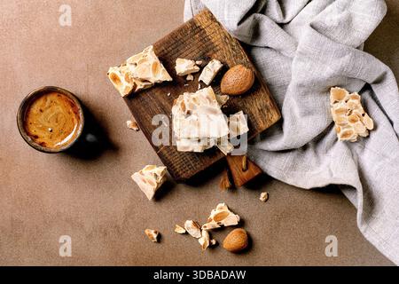 Dessert espagnol traditionnel turron croustillant aux amandes, cassé en morceaux sur une planche de service en bois. Serviette en lin, café expresso en tasse noire. Beige Banque D'Images