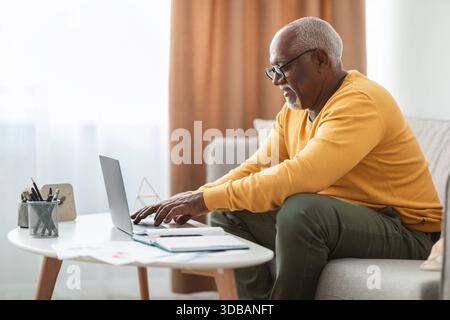 Homme senior travaillant sur ordinateur portable à la maison tout en étant assis sur le canapé dans le salon confortable pendant les heures de jour Banque D'Images