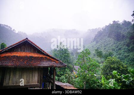 Maison traditionnelle en bois avec un toit de tuiles oranges nichée dans un paysage de montagne brumeux et verdoyant, évoquant une atmosphère rurale sereine. Banque D'Images