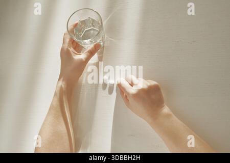 Mains de la personne se préparant à prendre des médicaments avec un verre d'eau sur la table blanche Banque D'Images