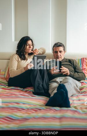 Couple d'âge moyen allongé sur le lit à la maison utilisant la technologie. Femme tenant une tablette numérique et homme utilisant un smartphone. Scen authentique style de vie domestique Banque D'Images