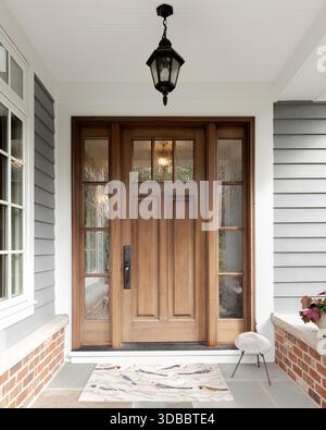 Une porte d'entrée et un détail de porche couvert sur une maison avec parement gris, garniture blanche, brique rouge, une porte d'entrée en bois. Banque D'Images