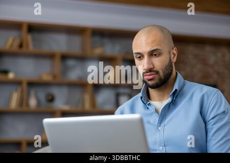 Jeune homme chauve se concentrant sur le travail tout en utilisant un ordinateur portable dans un environnement de bureau à domicile moderne avec une étagère en arrière-plan, se concentrant sur le travail à distance et l'utilisation d'Internet Banque D'Images