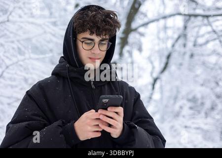 Portrait d'un adolescent concentré utilisant un smartphone à l'extérieur dans Snowy Winter Park. Jeune homme vérifiant les médias sociaux ou texto, portant des lunettes et PUF noir Banque D'Images