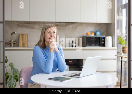 Femme âgée assise à une table dans une cuisine moderne, reposant son menton sur des mains serrées tout en regardant soigneusement loin de son ordinateur portable, représentant le travail à distance et la planification Banque D'Images