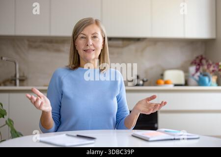Femme mature communiquant pendant un appel vidéo en ligne ou une réunion virtuelle depuis sa cuisine à domicile, faisant activement des gestes tout en ayant une discussion ou une conférence de travail à distance Banque D'Images