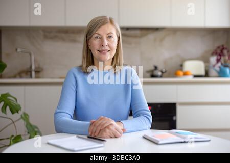 Femme mature assise à une table avec des cahiers, souriant avec confiance à la caméra tout en travaillant à distance ou en participant à un appel vidéo depuis sa cuisine moderne Banque D'Images