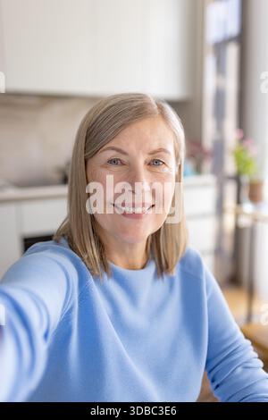 Femme âgée avec des cheveux blonds à la longueur de l'épaule posant pour un selfie ou ayant un appel vidéo amical tout en regardant la caméra et en souriant largement dans un cadre lumineux de cuisine à la maison Banque D'Images