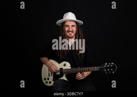 Un musicien jouant de la guitare électrique, portant un chapeau blanc et une chemise noire, avec de longs dreadlocks, sur un fond noir Banque D'Images