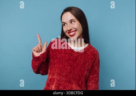 Une femme portant un pull rouge se tient sur un fond bleu. Elle sourit et pointe son doigt vers le haut avec une expression excitée tout en regardant l'AC Banque D'Images