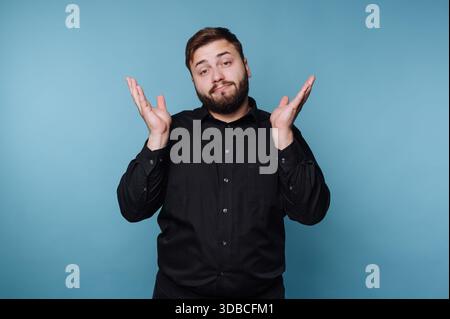 Un homme se tient debout les mains levées, montrant une expression confuse. Il porte une chemise noire et est dans un studio avec un fond bleu. Cette scène capture Banque D'Images