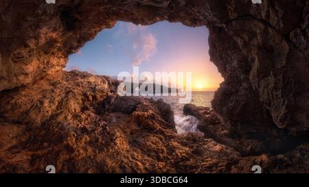 Vue de l'intérieur d'une grotte marine encadrant une côte rocheuse accidentée au lever du soleil, avec une lumière douce se reflétant sur la mer Méditerranée et les vagues se brisant Banque D'Images