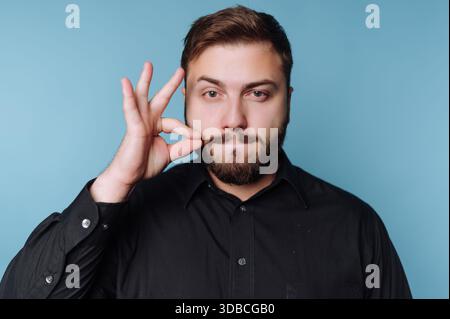 Un homme se tient sur un fond bleu clair. Il tient une main avec les doigts dans un geste précis tout en gardant une expression sérieuse sur son visage. Banque D'Images