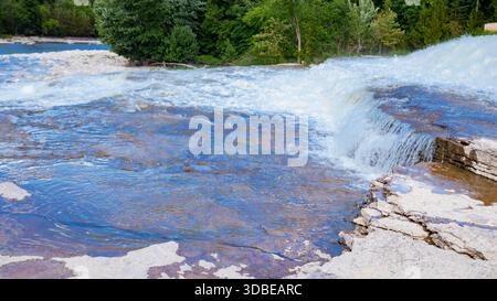 Une puissante cascade à plusieurs niveaux descend en cascade sur des formations rocheuses en couches dans un cadre forestier luxuriant. Les courants blancs mousseux contrastent avec le ciel dense. Banque D'Images