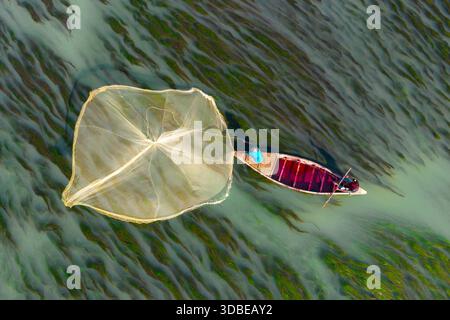 Natore, Bangladesh - 13 décembre 2025 : vue aérienne d'un pêcheur solitaire sur un bateau en bois, jetant un filet dans l'eau tourbillonnante et striée d'algues de la rivière Banque D'Images