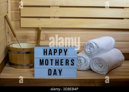 Un cadre confortable de sauna avec un panneau qui indique « Happy Mothers Day ». Il y a des serviettes blanches soigneusement roulées et un seau en bois. Banque D'Images