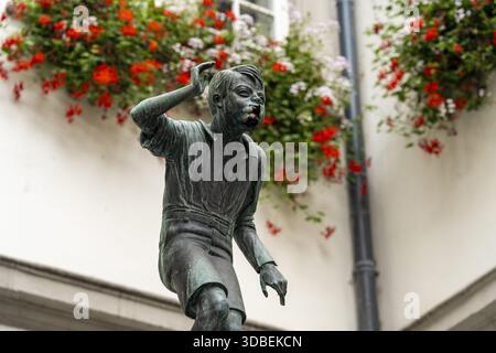 Vue d'une statue en bronze d'un garçon se grattant la tête sous des géraniums rouges vibrants en cascade d'un bâtiment, un mélange d'art et de nature, Coblence, Rhinel Banque D'Images