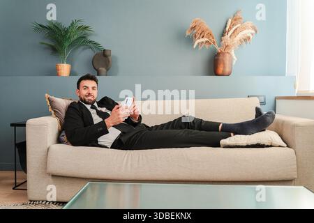 Un homme d'affaires détendu est confortablement allongé sur un canapé confortable tout en utilisant son smartphone. Il semble engagé et concentré, mettant en valeur un mélange de loisirs Banque D'Images