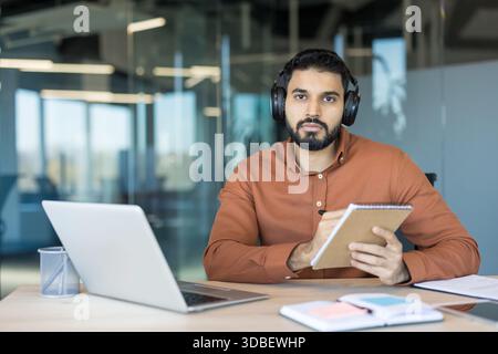 Jeune homme portant des écouteurs et tenant un bloc-notes, regardant la caméra tout en travaillant ou étudiant à distance à un bureau dans un environnement de bureau contemporain Banque D'Images