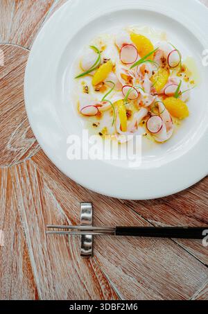Le ceviche de poisson est disposé sur une plaque circulaire en céramique garnie de radis et de ciboulette. Une sauce jaune-orange scintille sur la protéine de poisson blanc al Banque D'Images