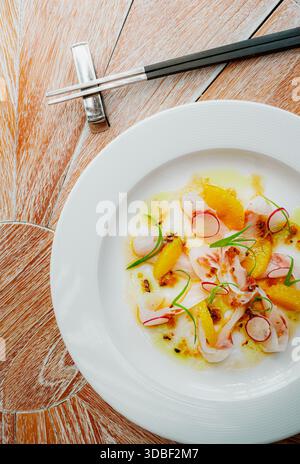 Le ceviche de poisson, composé de tranches de poisson cru, de segments d'agrumes vibrants et de radis finement tranchés, est habilement disposé sur une assiette blanche avec une sauce crémeuse, un Banque D'Images