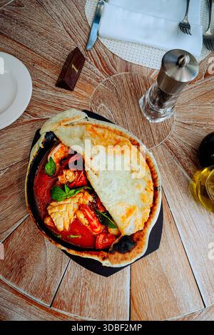 Un ragoût de fruits de mer en croute présente des calmars, des crevettes et du crabe sur une assiette en céramique dans un plat en métal. Feuilles de basilic et fromage garnissent le plat, accompagnie Banque D'Images