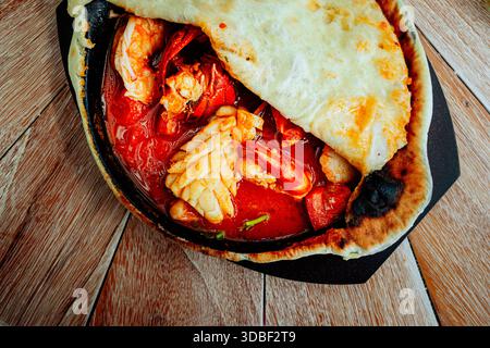 Un ragoût de fruits de mer en croute, composé de crevettes, de calmars et de crabe, mijote dans un récipient de cuisson sombre au sommet d'une surface rustique en bois. Tomates et un rouge visqueux Banque D'Images