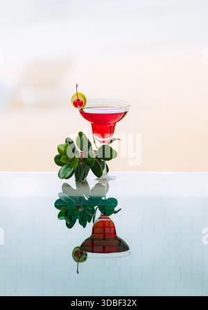 Un cocktail vibrant avec une cerise maraschino et des feuilles verdoyantes repose dans un verre élégant. Une surface en miroir améliore l'attrait de la boisson contre un SAF Banque D'Images