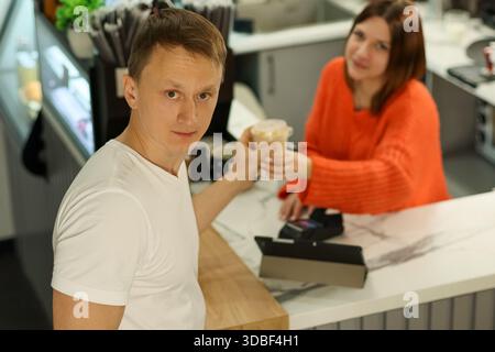Jeune patron masculin regarde la serveuse en orange comme elle lui remet une boisson crémeuse sur le comptoir de marbre du café Banque D'Images