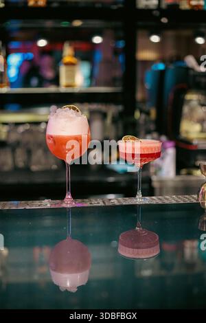 Vue rapprochée élégante de deux cocktails rouges dans des verres à pied sur un comptoir de bar moderne, avec mousse, garniture d'agrumes et ambiance nocturne trépidante Banque D'Images