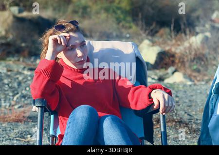 Femelle en col roulé rouge touchant des lunettes sur la tête tout en se prélassant sur une chaise pliante près de la rive rocheuse au soleil. Banque D'Images