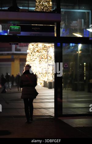 Silhouette d'une personne marchant à travers des portes vitrées vers une rue de ville illuminée par des lumières festives la nuit à Katowice, Pologne Banque D'Images